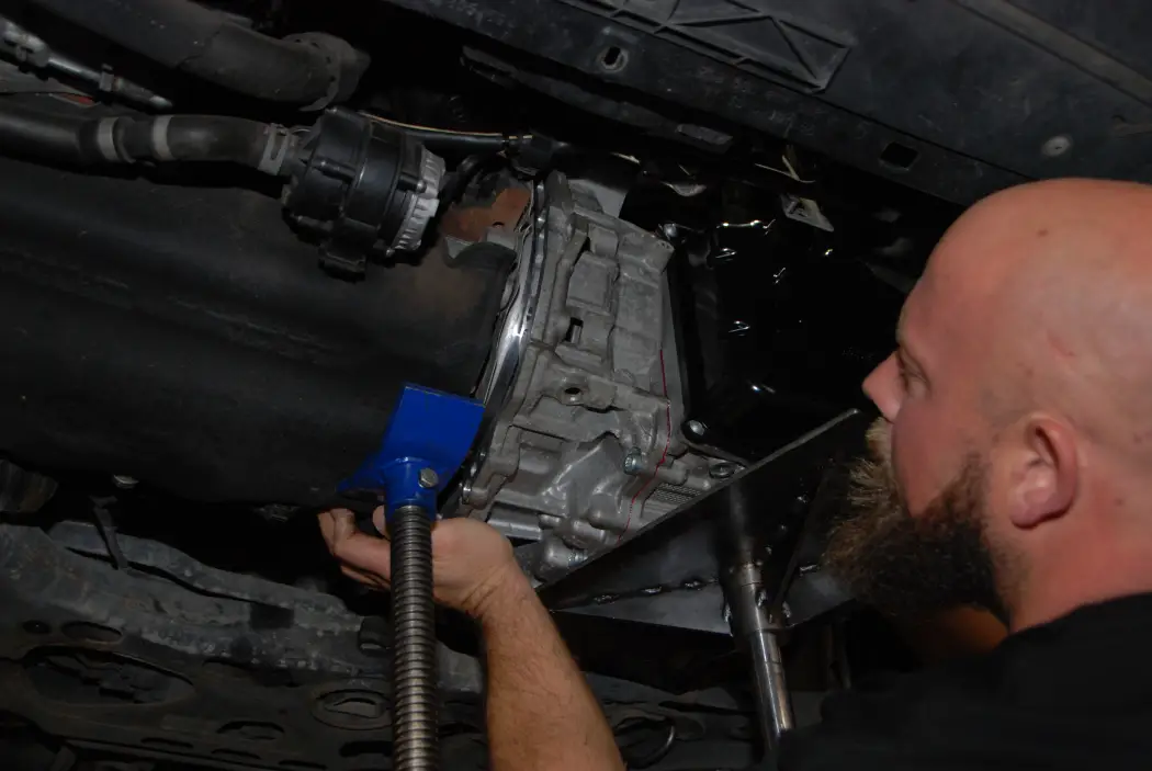 A mechanic named Arron removing an automatic transmission from a vehicle for rebuild, using a transmission jack to support the unit while working underneath the car in a workshop.