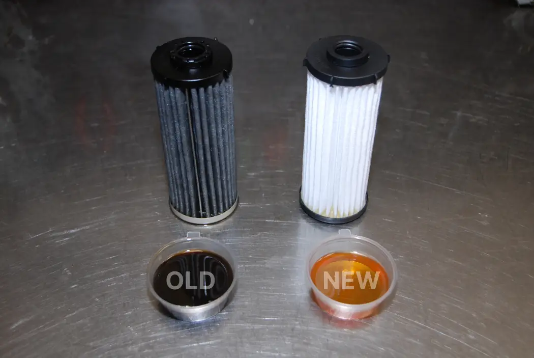 Comparison of old and new DSG filters and transmission fluid, showing a dark used filter and oil sample labeled 'OLD' on the left, and a clean white filter with bright amber fluid labeled 'NEW' on the right, placed on a metal workbench.